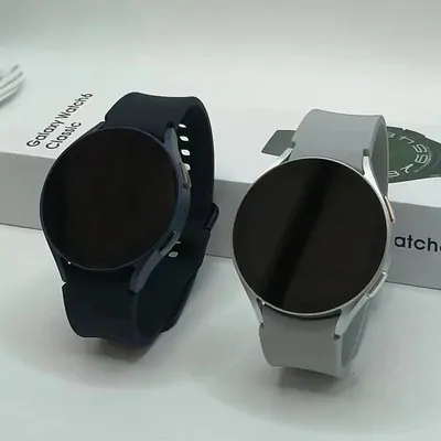 Two round smartwatches, one on a black strap and one on a grey strap, standing beside their boxes
